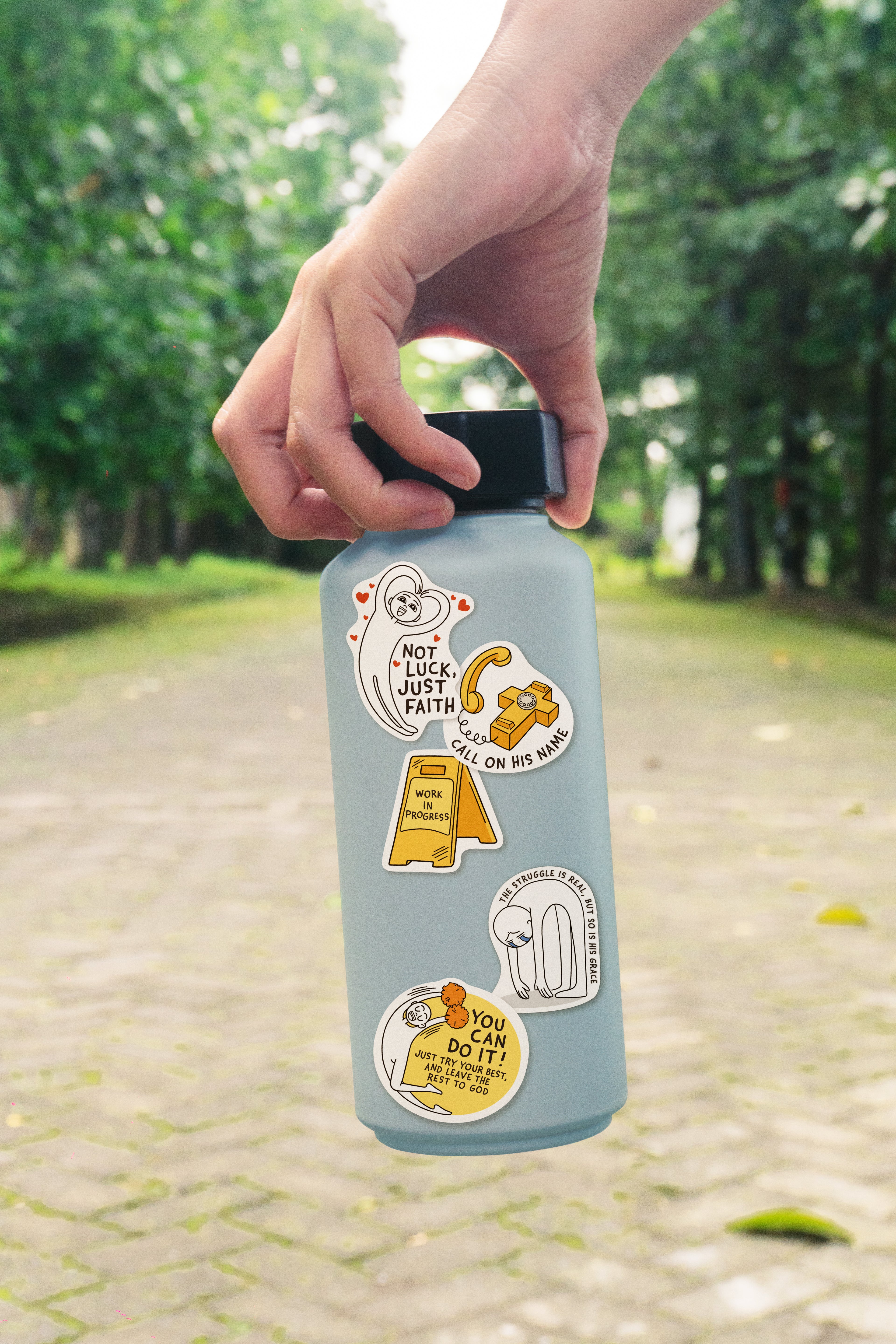 Hand holding a water bottle decorated with Everyday Believer stickers featuring Christian messages like "Not Luck, Just Faith" and "Call on His Name," from The Commandment Co, Singapore.