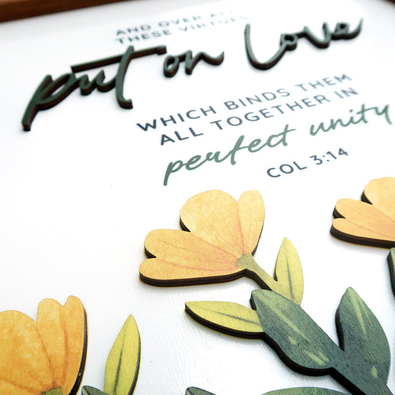 Close-up of a wooden scripture poster featuring the text "Put on Love" from Colossians 3:14. Includes layered lettering and decorative yellow flowers with green leaves.
