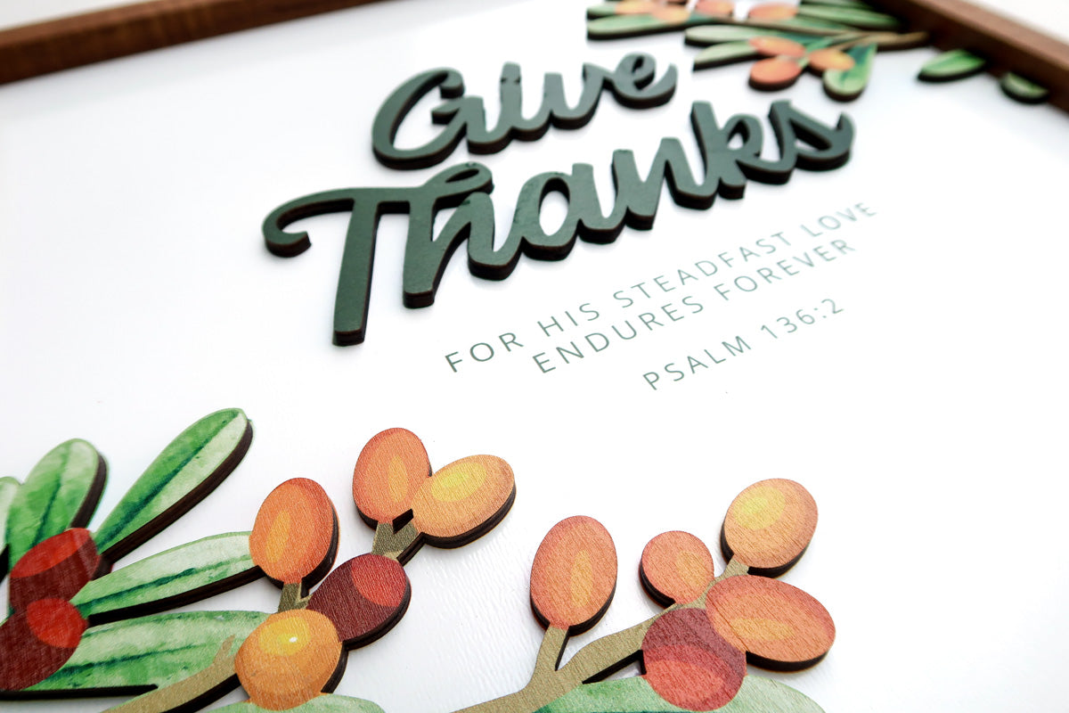 Close-up of a handcrafted wooden poster with the text "Give Thanks" and scripture from Psalm 136:2. Features layered text and decorative orange berries with green leaves.