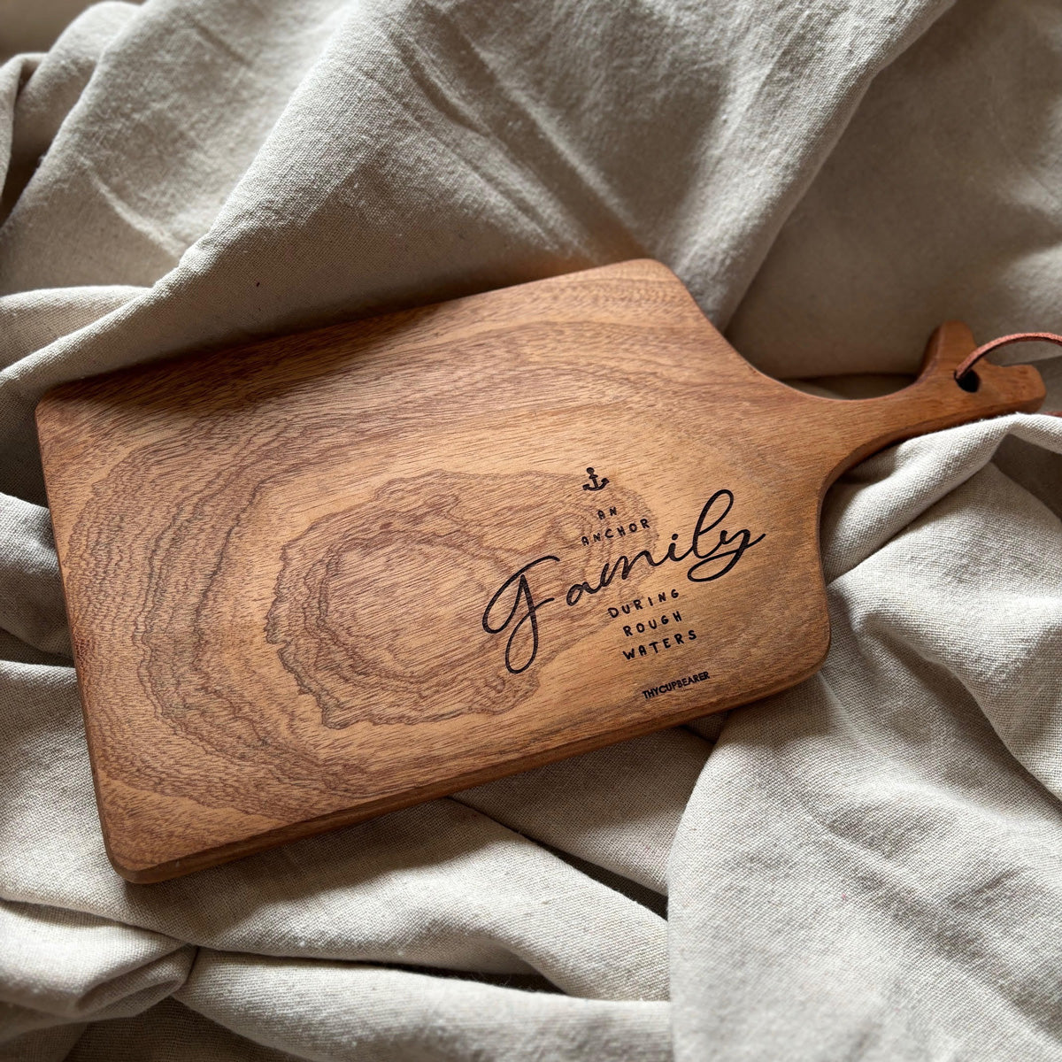 A wooden cheese board engraved with the phrase "Family, an anchor during rough waters," featuring an anchor symbol. The board showcases a natural wood grain pattern, styled on a soft beige fabric. A perfect rustic kitchen decor piece or thoughtful family-oriented gift.