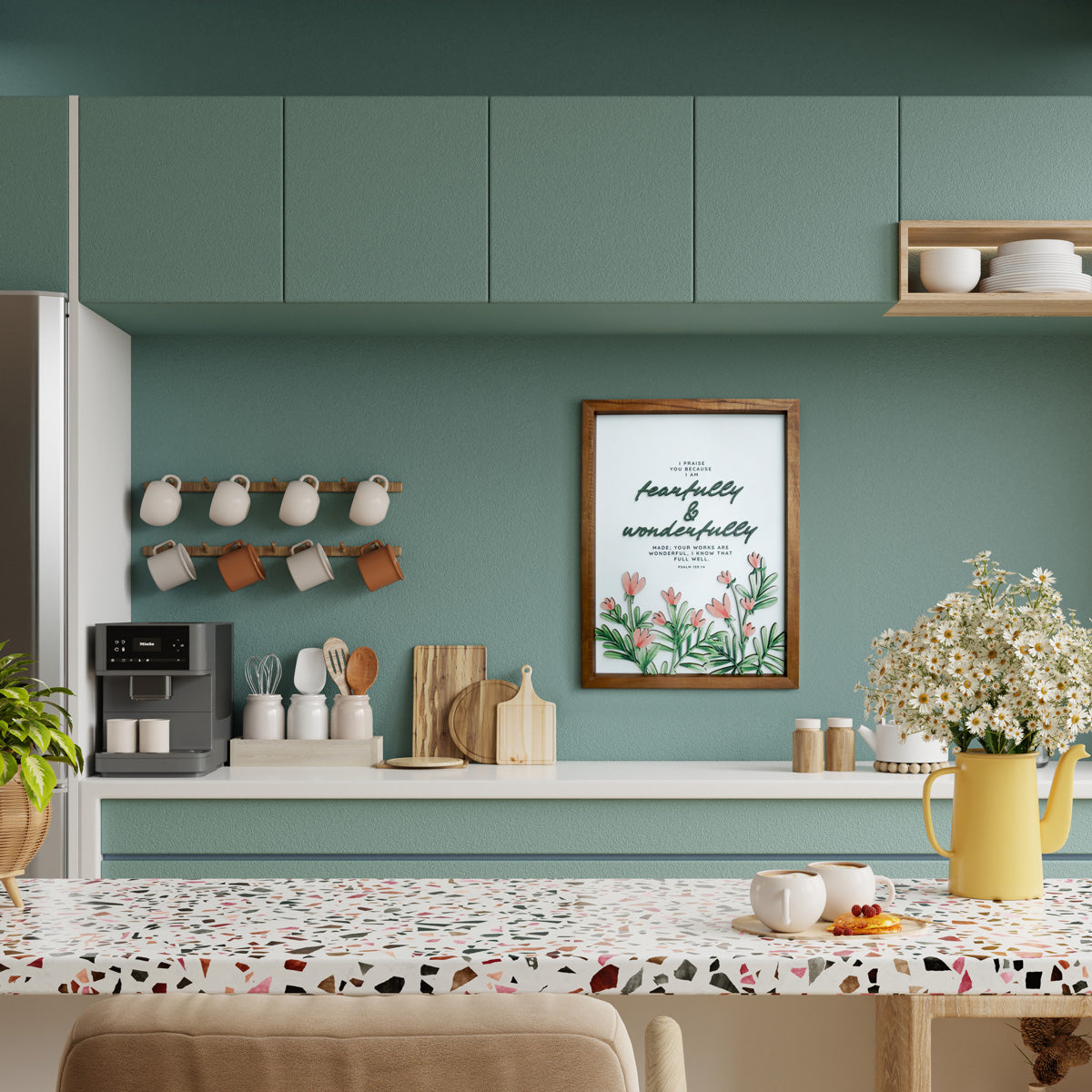 A wooden-framed wall art featuring the Bible verse "Fearfully & Wonderfully Made" from Psalm 139:14. The artwork is decorated with delicate pink flowers and green foliage, adding a calming and inspirational touch to the modern kitchen. The setting includes a terrazzo-patterned countertop, neatly arranged kitchenware, and a yellow pitcher filled with white flowers, creating a warm and inviting atmosphere perfect for Christian home decor.