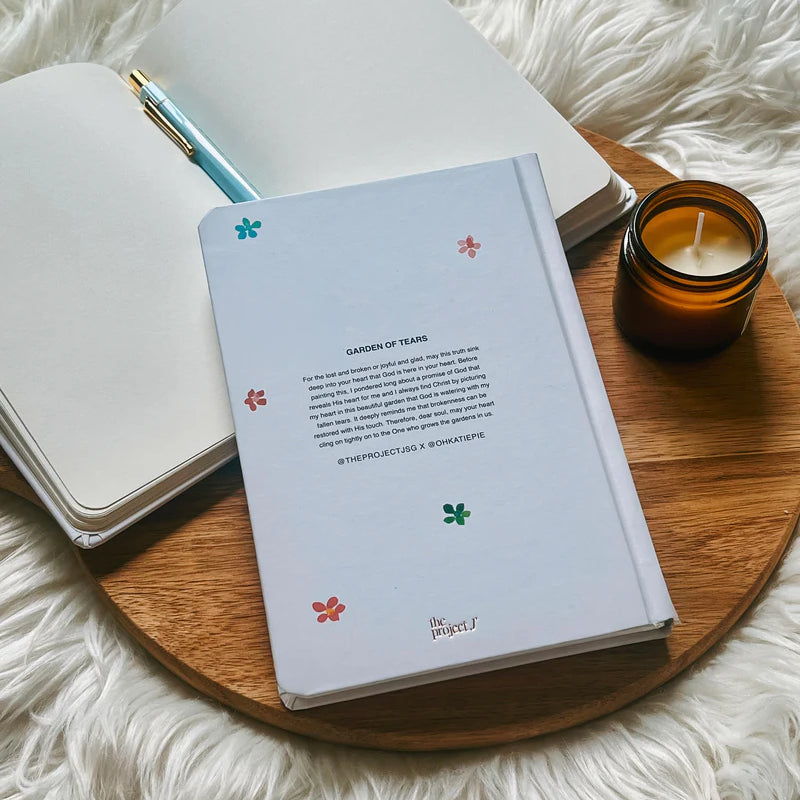 Off-white hard cover journal with printed message on back cover by Project J placed on wooden tray beside open notebook and candle.