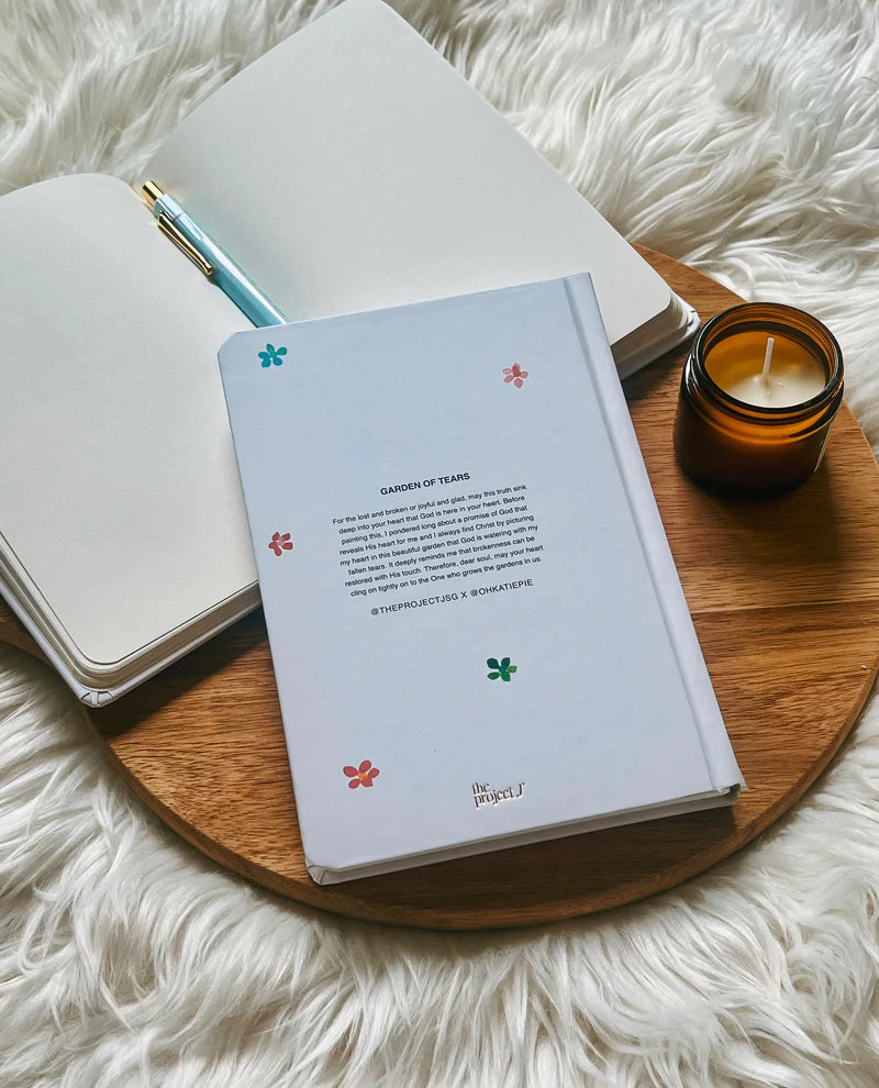 Off-white hard cover journal with printed message on back cover by Project J placed on wooden tray beside open notebook and candle.