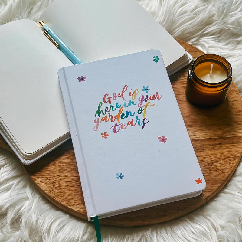 Off-white hard cover journal with “God is here in your garden of tears” text design by Project J placed on wooden tray beside open notebook and candle.