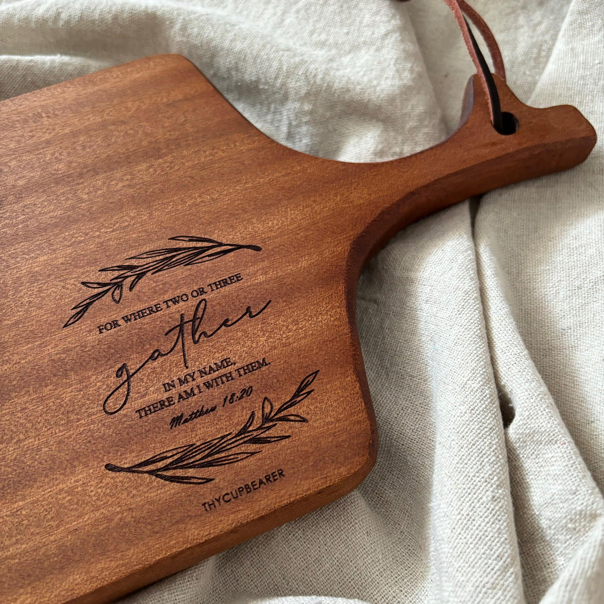 A rustic wooden cheese board engraved with the Bible verse "For where two or three gather in My name, there am I with them" (Matthew 18:20), surrounded by decorative olive branches. The board features a leather strap and is displayed on a soft beige fabric, making it a perfect kitchen decor item or thoughtful Christian gift.
