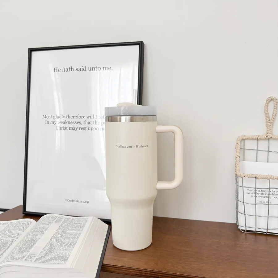 Cream insulated cup with the words "God has you in His heart" on a wooden table, surrounded by a Bible and inspirational Christian decor, reflecting simple happiness and encouragement from Jincha God.