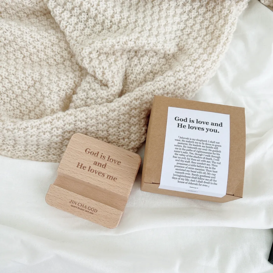 Wooden phone holder engraved with "God is love and He loves me," displayed next to its box with an encouraging message, on a cozy knitted blanket, inspiring faith and simple happiness.