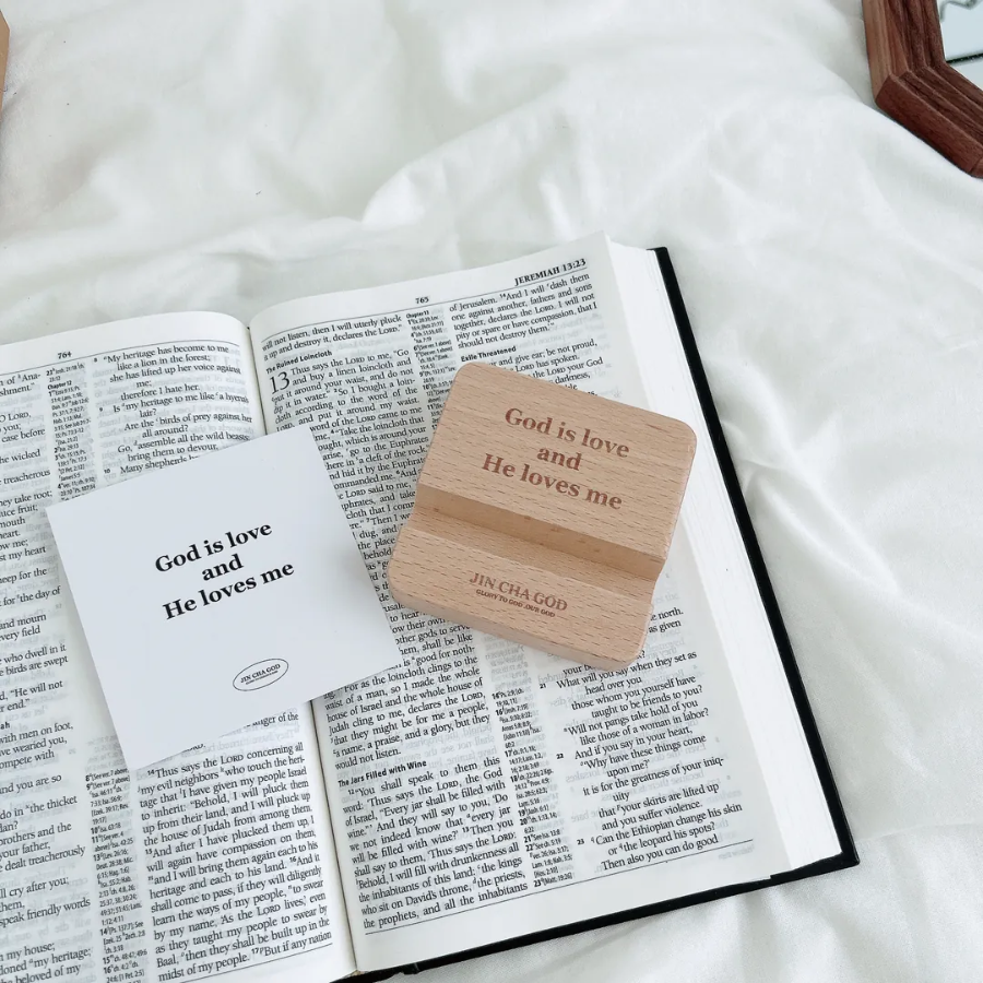 Wooden phone holder engraved with the message "God is love and He loves me," displayed on an open Bible with an inspirational card, encouraging faith and simple happiness.
