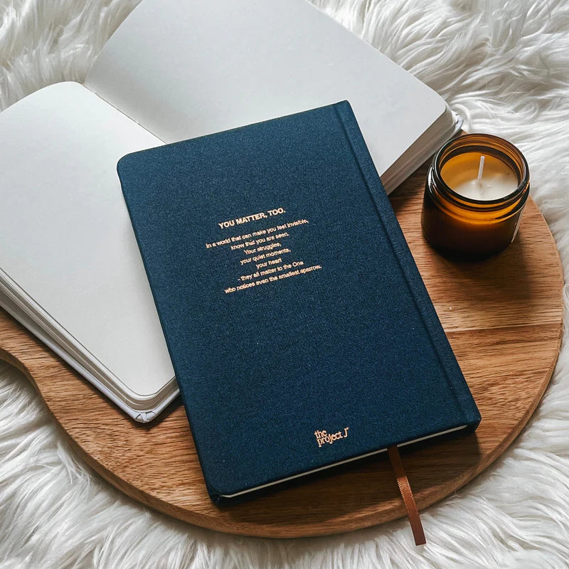 Blue fabric journal with gold text design on back cover by Project J placed on wooden tray beside open notebook and candle.
