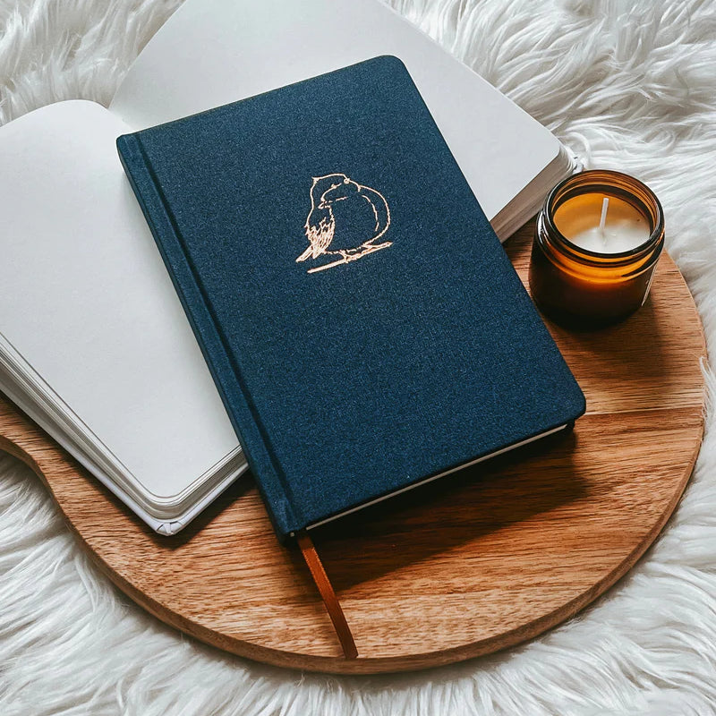 Blue fabric hardcover journal by Project J with gold bird design, placed on wooden tray beside open notebook and candle.
