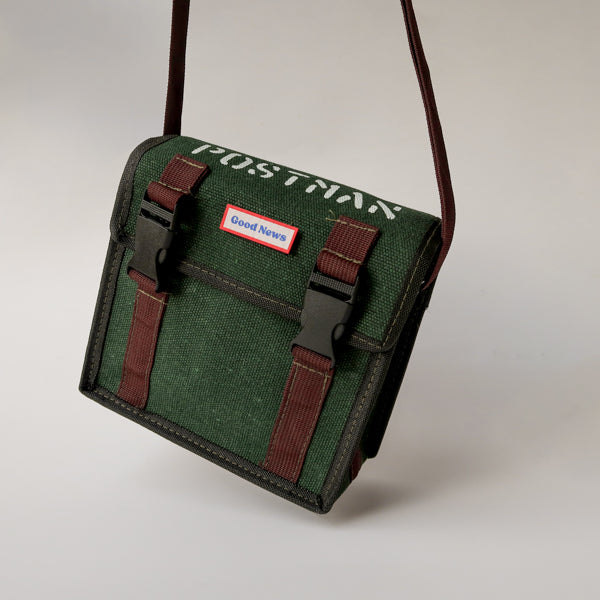 A green canvas messenger bag with 'Good News' branding, featuring brown straps and sturdy clips. The bag has a faith-inspired design and is ideal for daily use, with a minimalist style and practical functionality.