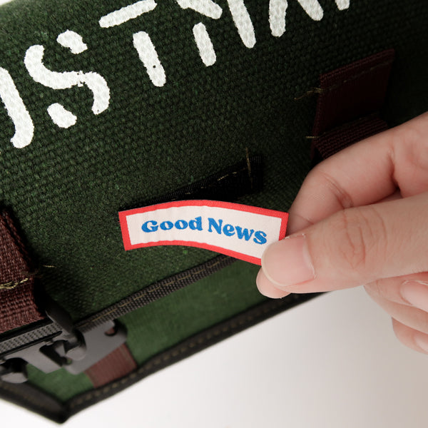 A close-up view of a green canvas messenger bag with the label 'Good News' being attached. The bag has 'POSTMAN' text on the top and a faith-based theme, showcasing both practicality and inspiration.