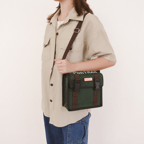 A person wearing a green canvas messenger bag with 'Good News' branding, shown from the side with the bag worn crossbody. The bag features brown straps and clips, offering a practical and faith-inspired design.