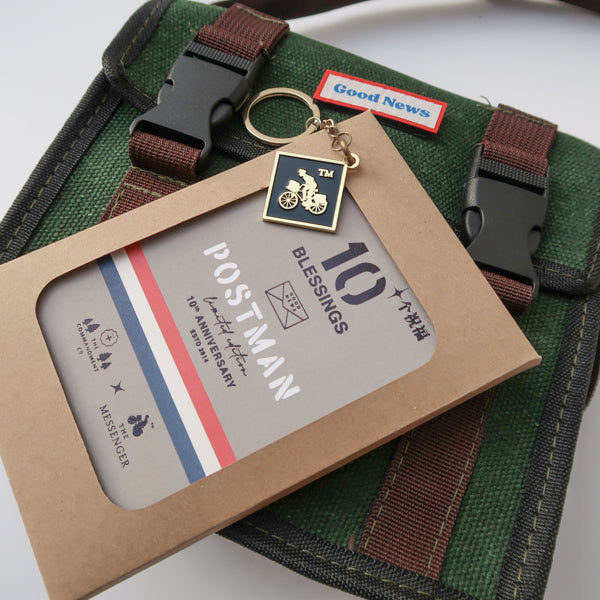 Close-up of a green canvas 'Good News' messenger bag with 'POSTMAN' text, displayed with a 10th anniversary edition 'Postman' keychain and packaging. The bag’s brown straps complement the faith-inspired design.