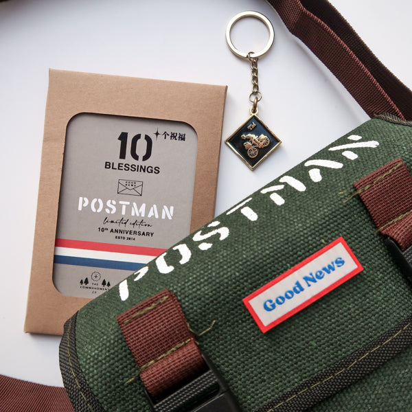 A green canvas 'Good News' messenger bag with brown straps displayed alongside the 10th anniversary edition 'Postman' keychain and packaging. The design features a bold 'POSTMAN' label on the bag and a small motorbike-themed keychain.