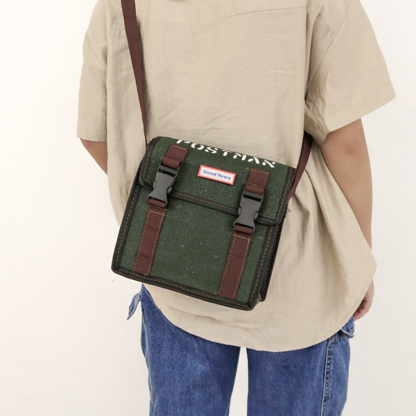 A rear view of a person wearing a green canvas messenger bag with 'Good News' branding. The bag is faith-themed with 'POSTMAN' text on top and brown straps, suitable for everyday use.