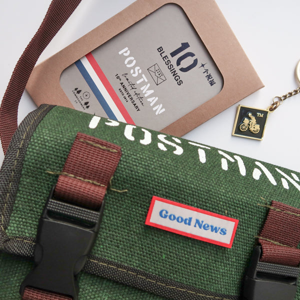 A close-up of a green canvas messenger bag with 'Good News' branding, featuring a 10th anniversary edition 'Postman' keychain and packaging. The brown straps and keychain complement the faith-inspired design.