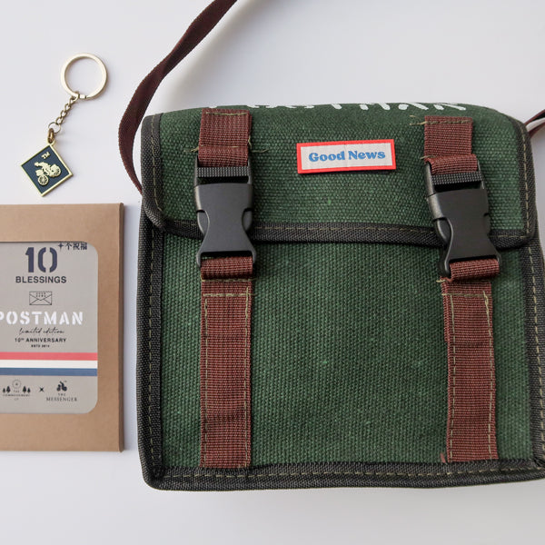 A green canvas messenger bag with 'Good News' branding displayed alongside the 10th anniversary 'Postman' keychain and packaging. The bag has brown straps and a faith-themed design, symbolizing both practicality and inspiration.