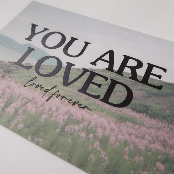 A detailed view of a postcard with a scenic field of pink flowers and the heartfelt message "You Are Loved, loved forever," offering reassurance and comfort.