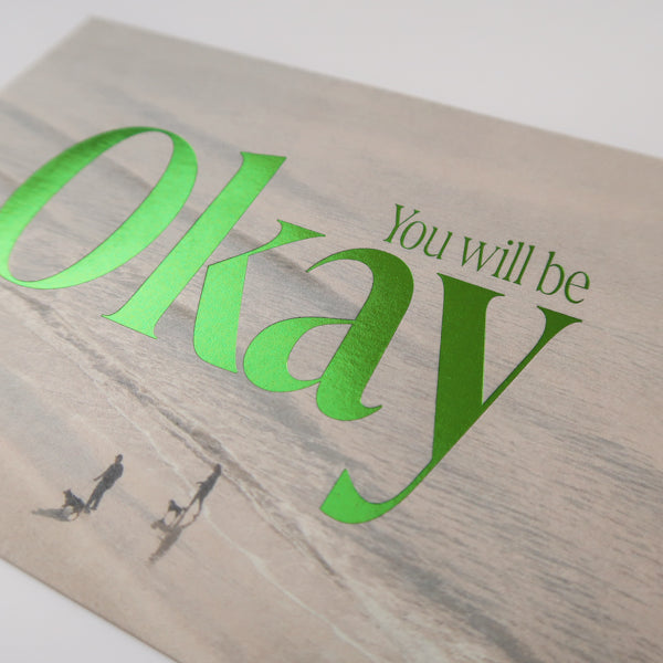 A detailed view of a postcard with a serene beach background and the comforting text "You Will Be Okay" in bold green lettering, designed to offer reassurance and encouragement.