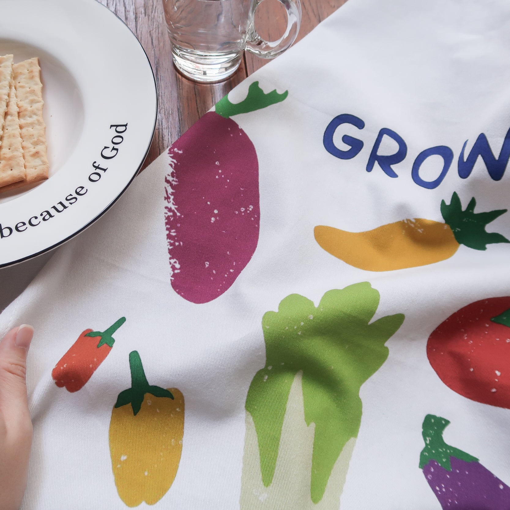 Grow in Grace tea towel with a colorful vegetable print on white fabric, shown beside a plate of crackers on a wooden table—a contemporary Christian kitchen decor gift.