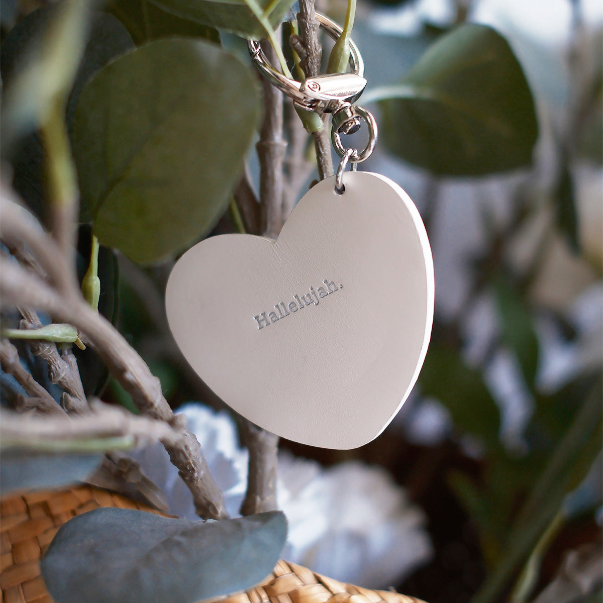 Heart-shaped keychain with 'Hallelujah' engraved in gold, hanging on a tree branch with green leaves in the background, symbolizing faith and inspiration.