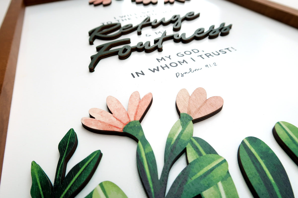 Close-up of a handcrafted wooden scripture poster featuring the text "Refuge and Fortress" from Psalm 91:2. Includes layered lettering and decorative pink flowers with green leaves.