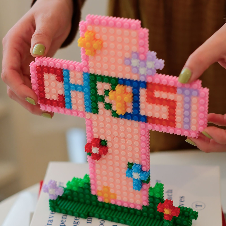 Hands holding a colorful micro-brick Christian cross with “CHRIST” lettering and flower accents, faith-inspired DIY decor from The Commandment Co