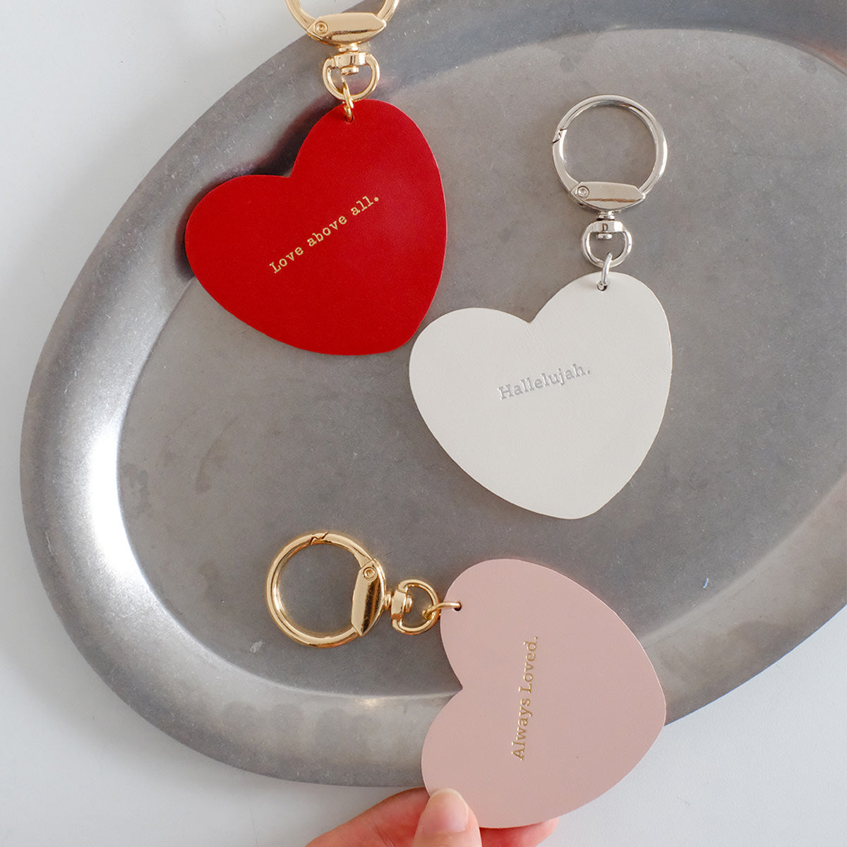 Three heart-shaped keychains with inspirational words: red with 'Love above all,' white with 'Hallelujah,' and pink with 'Always Loved,' displayed on a metallic tray.