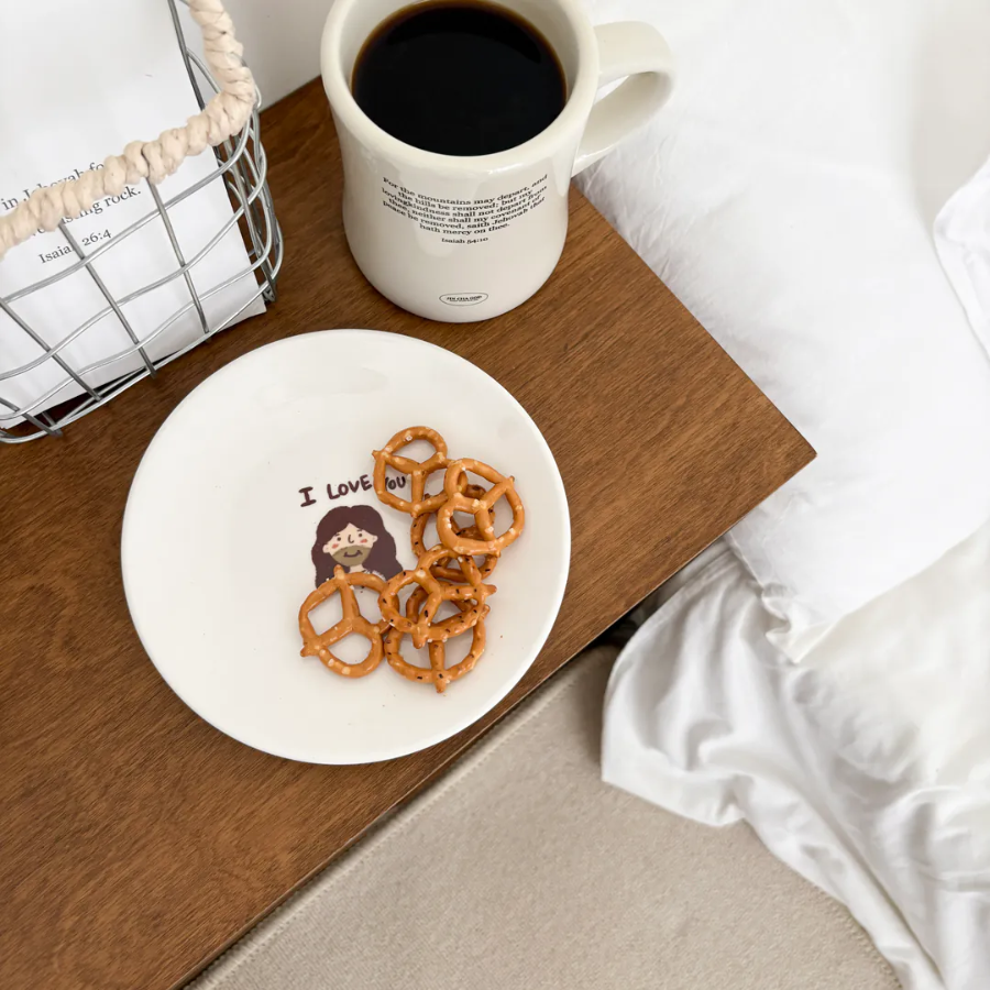 I Love You" dessert plate with a cute illustration of Jesus and pretzels, placed on a wooden bedside table next to a coffee mug, inspiring love and simple happiness as a Christian gift from Jincha God.