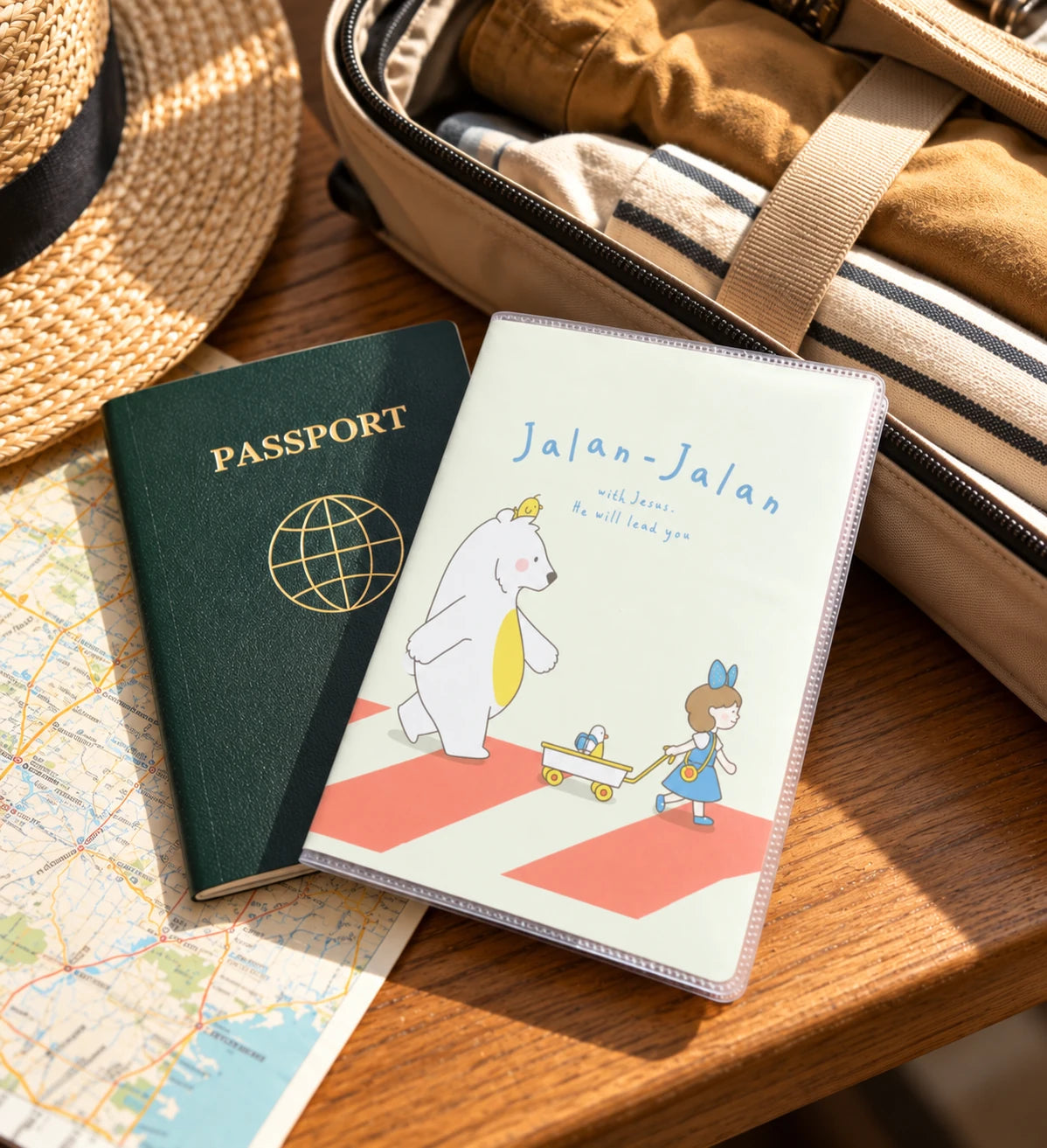 Jalan-Jalan With Jesus illustrated passport cover beside a passport, open luggage and travel map on a wooden table