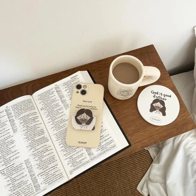 Hand-drawn Jesus phone grip used on a phone beside an open Bible, mug and Christian coaster in a cosy desk setting
