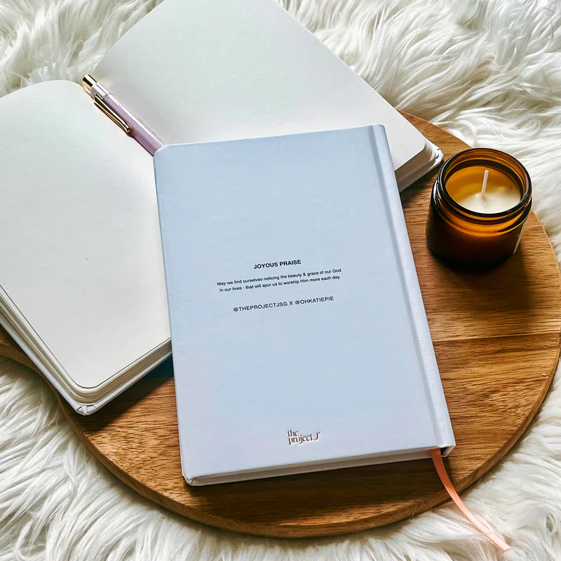 Off-white hard cover journal with printed message on back cover by Project J placed on wooden tray beside open notebook and candle.