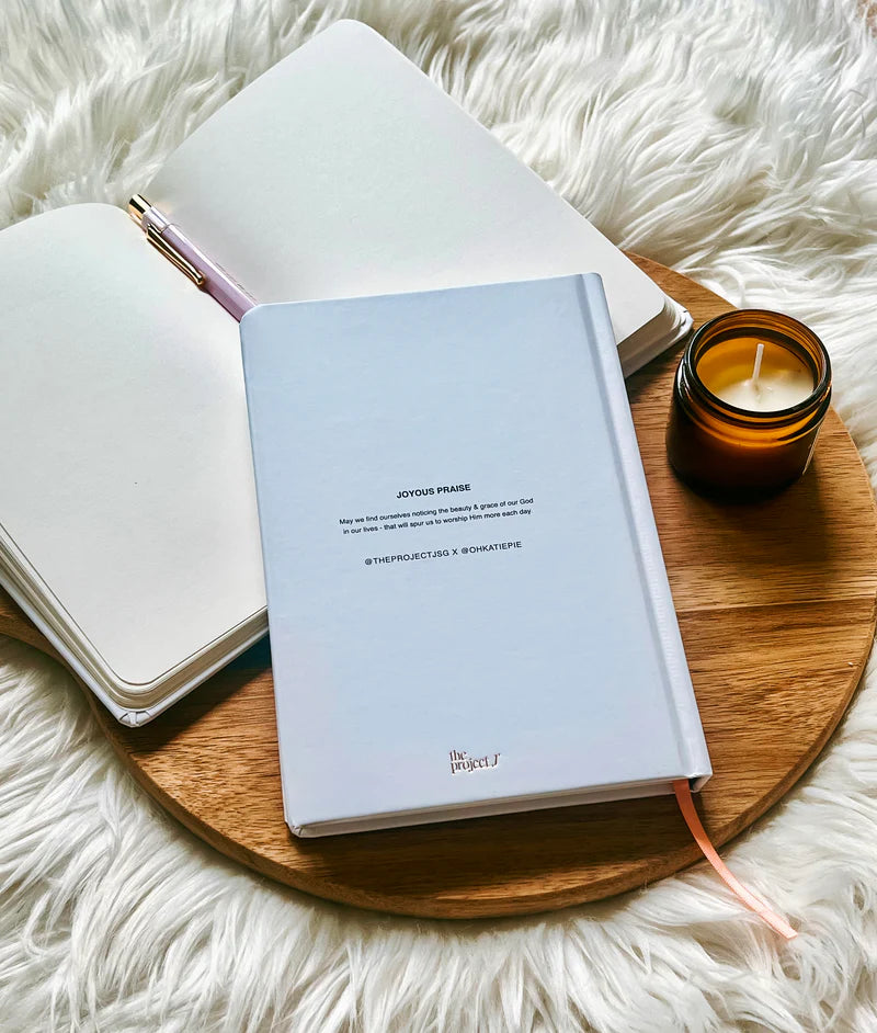 Off-white hard cover journal with printed message on back cover by Project J placed on wooden tray beside open notebook and candle.