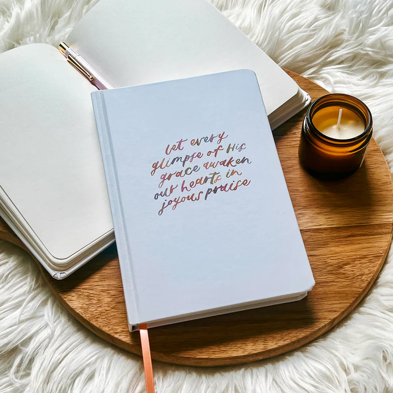 Off-white hard cover journal with “let every glimpse of His grace awaken our hearts in joyous praise” text by Project J placed on wooden tray beside open notebook and candle.