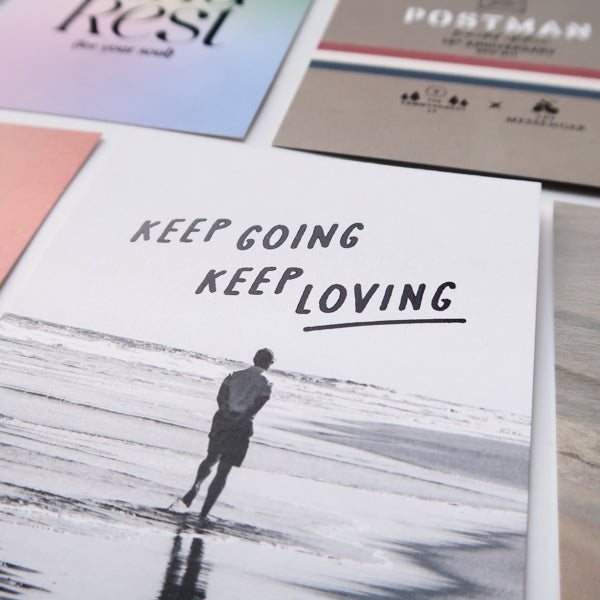 A close-up view of a collection of postcards, with a prominent black-and-white card featuring a person walking on the beach and the motivational message "Keep Going, Keep Loving" along with other faith-based and inspirational designs.