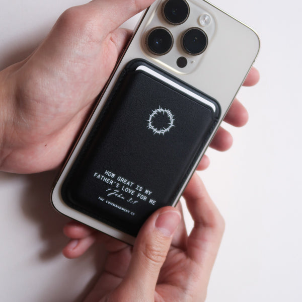 A pair of hands holding a mobile phone with the black Leather Magsafe Wallet attached to the back
