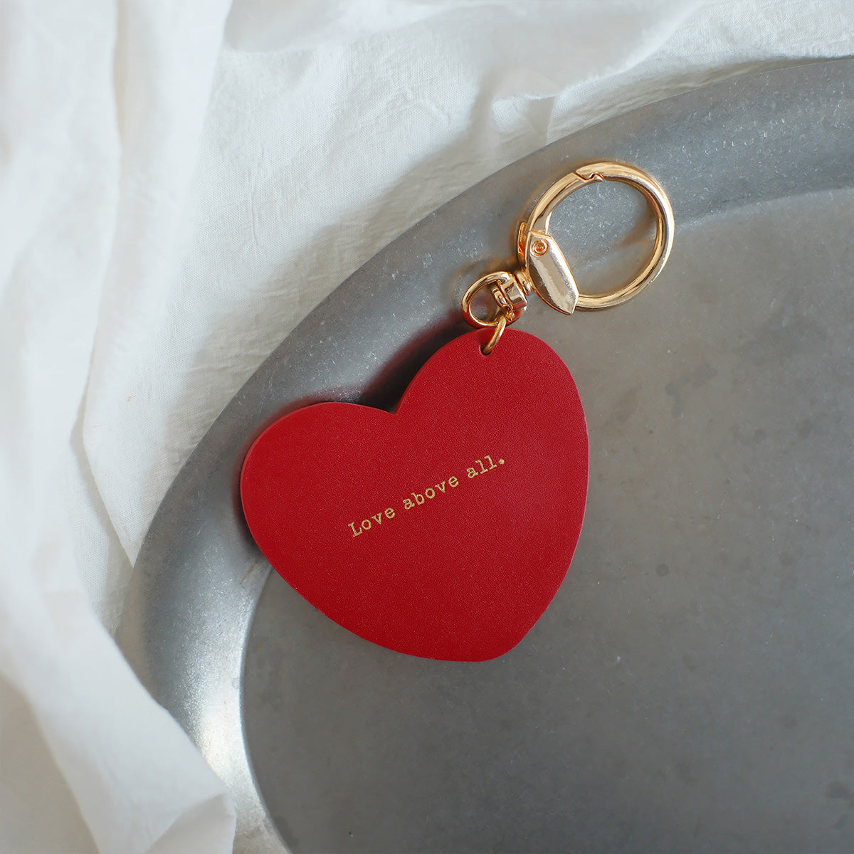 Heart-shaped red keychain engraved with 'Love above all,' a minimalist and faith-inspired accessory, perfect as a unique gift.