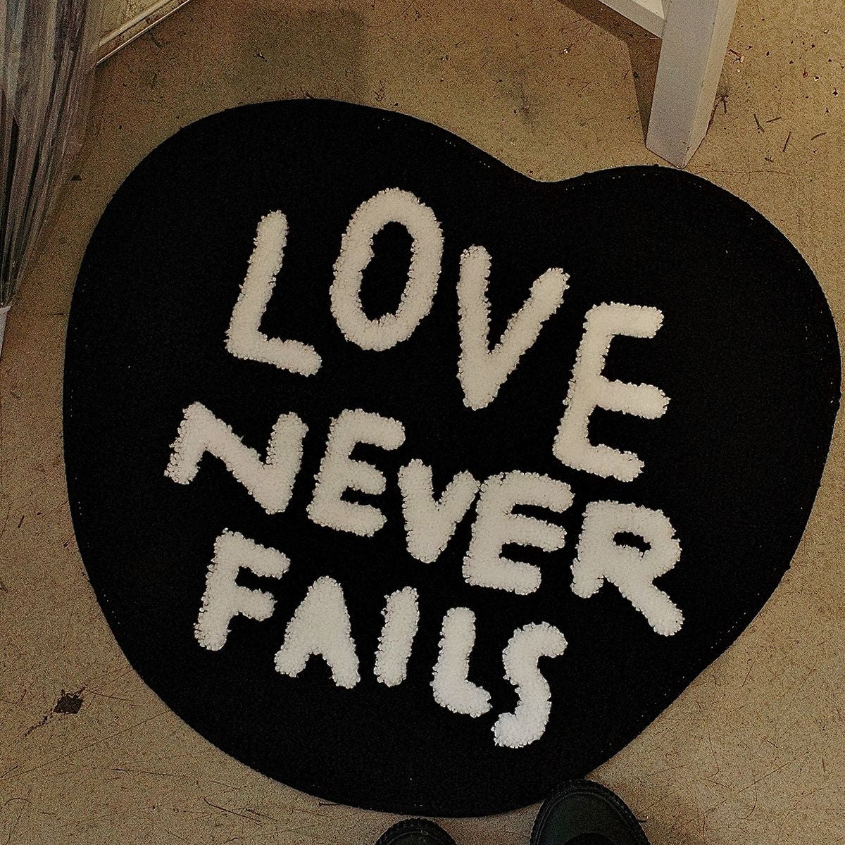 Black heart-shaped tufted rug with “Love Never Fails” lettering styled as Christian home decor indoors.