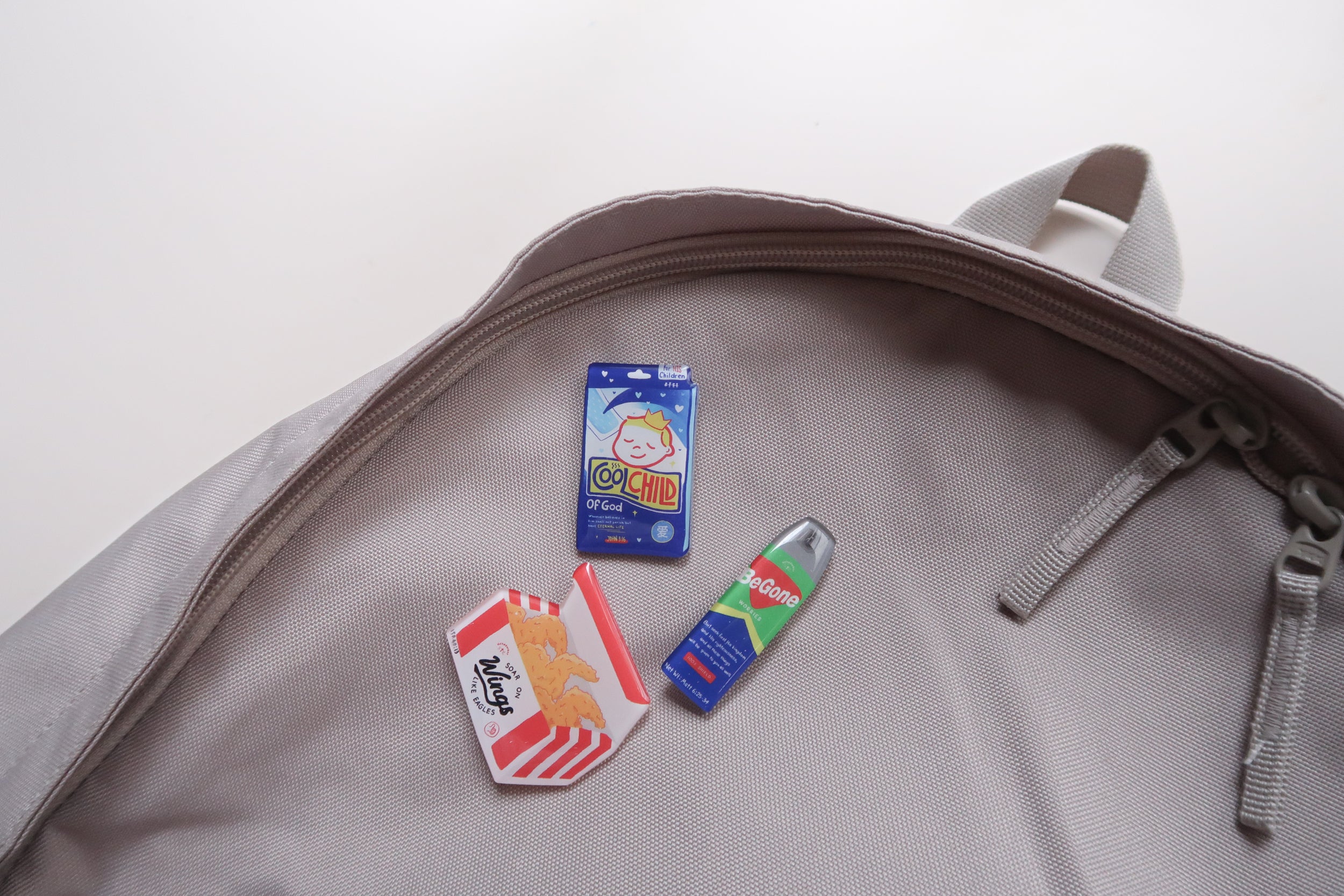 Love Supermarket acrylic pins featuring faith-inspired snack designs arranged inside a grey backpack, including Cool Child of God, BeGone worries spray, and Soar on Wings pin.