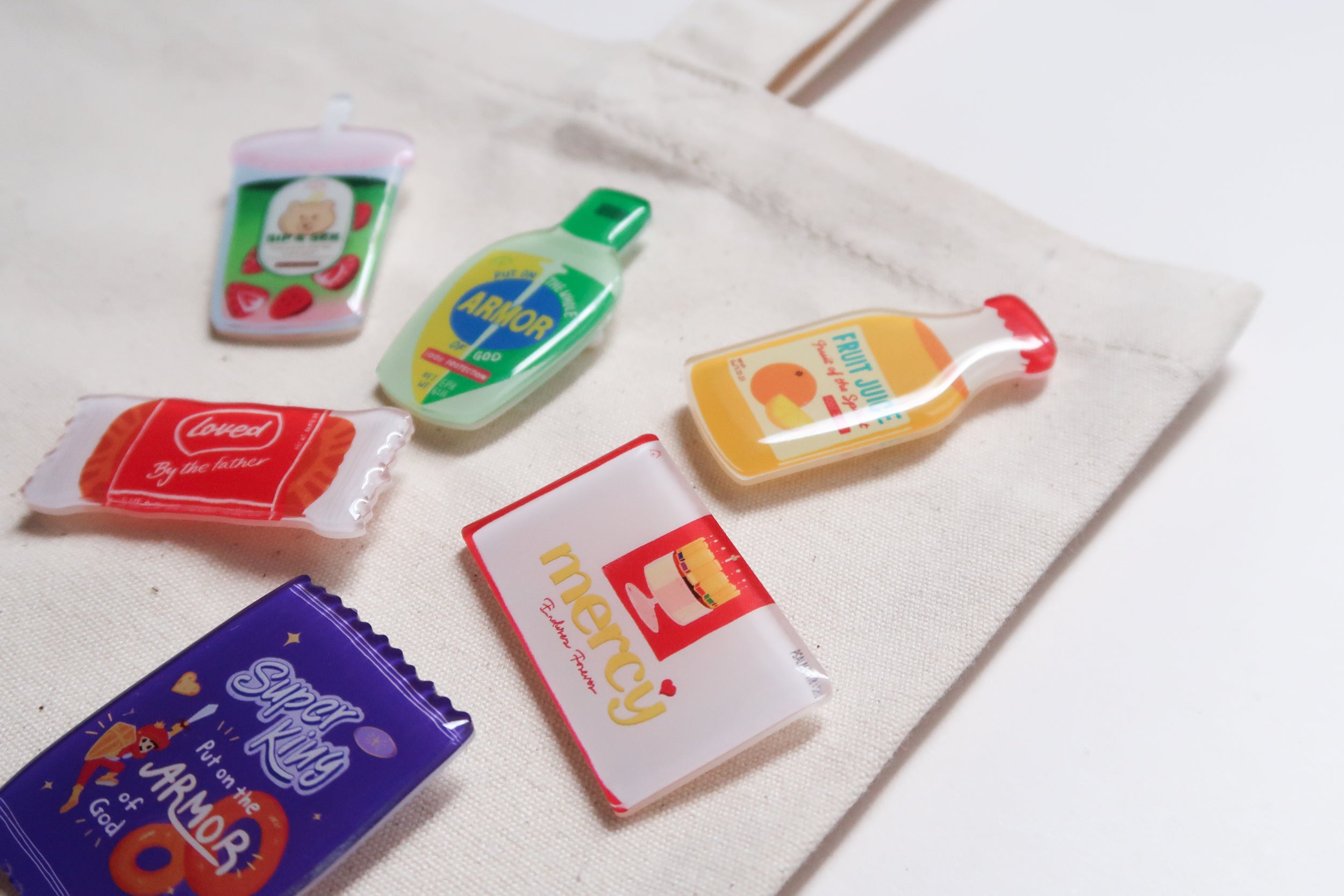 Love Supermarket acrylic pins styled on a natural canvas tote bag, featuring playful Christian designs inspired by Bible verses and everyday snacks.