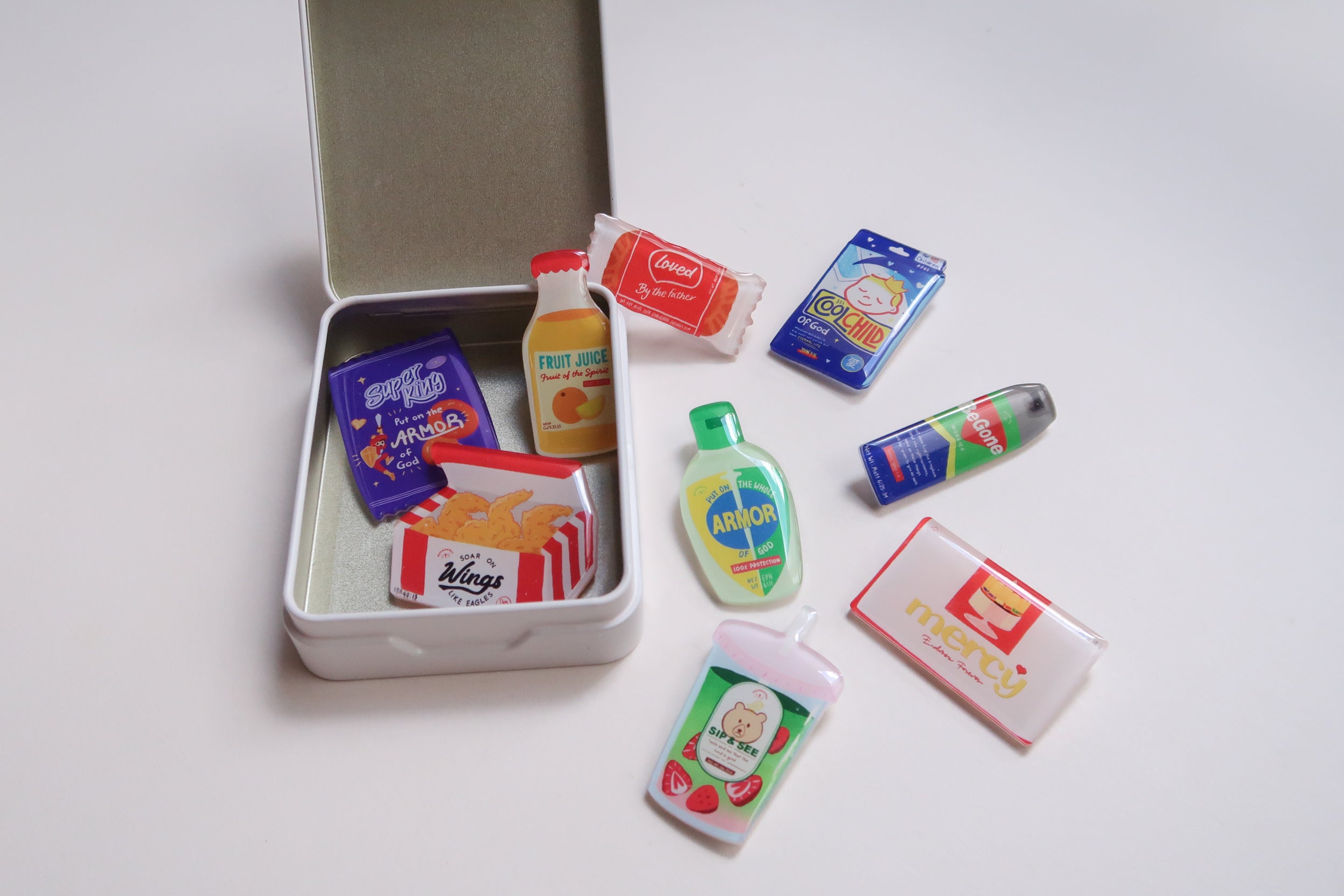 Love Supermarket acrylic pins displayed in and around a white tin box, showcasing Christian snack-themed designs like Armor of God, Fruit of the Spirit juice, Mercy chocolate, and Loved by the Father candy.