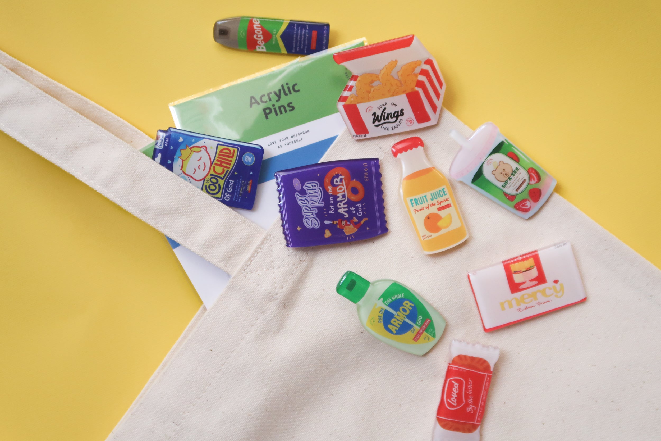 ove Supermarket acrylic pins arranged on a canvas tote against a yellow background, featuring colorful Christian snack-inspired designs from The Commandment Co.