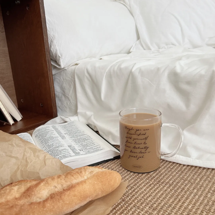 Clear glass mug with the message "Maybe you are dissatisfied with yourself from time to time. Actually, you have done a great job," filled with coffee and placed beside an open Bible and fresh bread, inspiring encouragement and simple happiness.