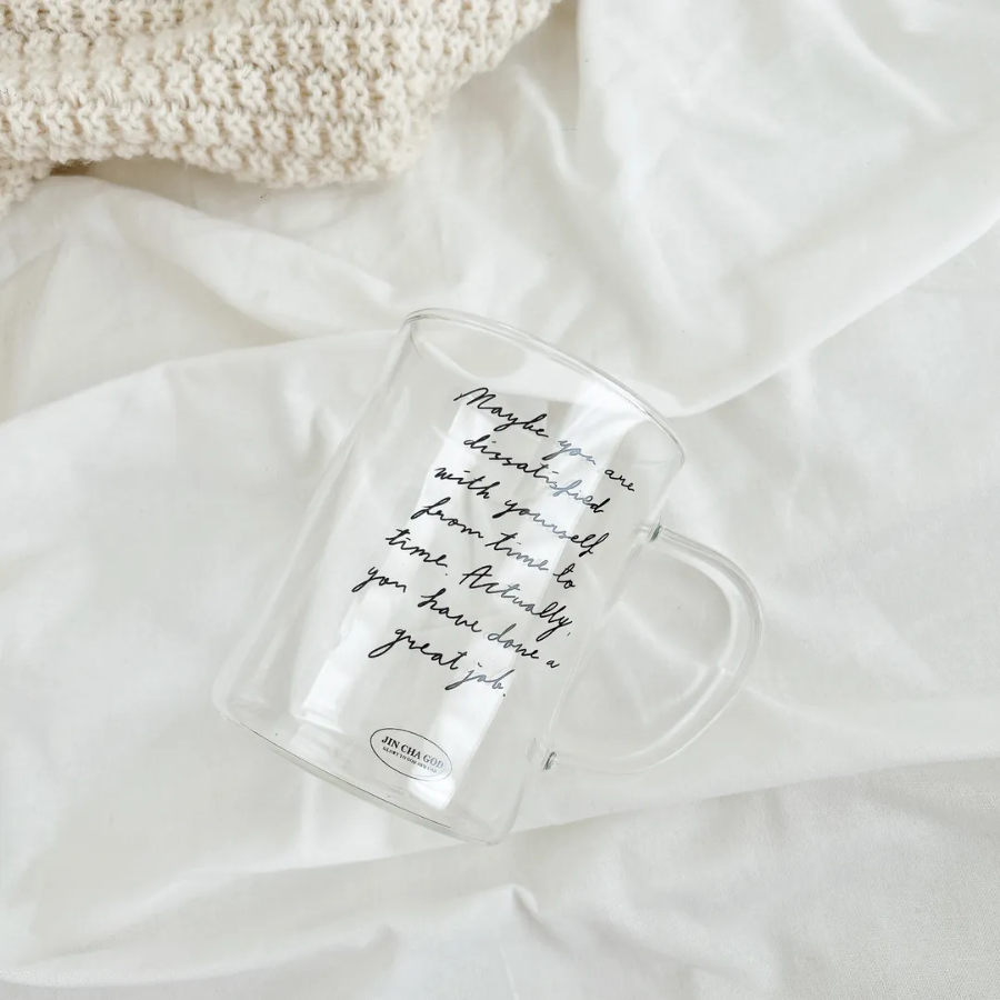 Clear glass mug with the handwritten message "Maybe you are dissatisfied with yourself from time to time. Actually, you have done a great job," placed on a soft white blanket, inspiring encouragement and simple happiness.
