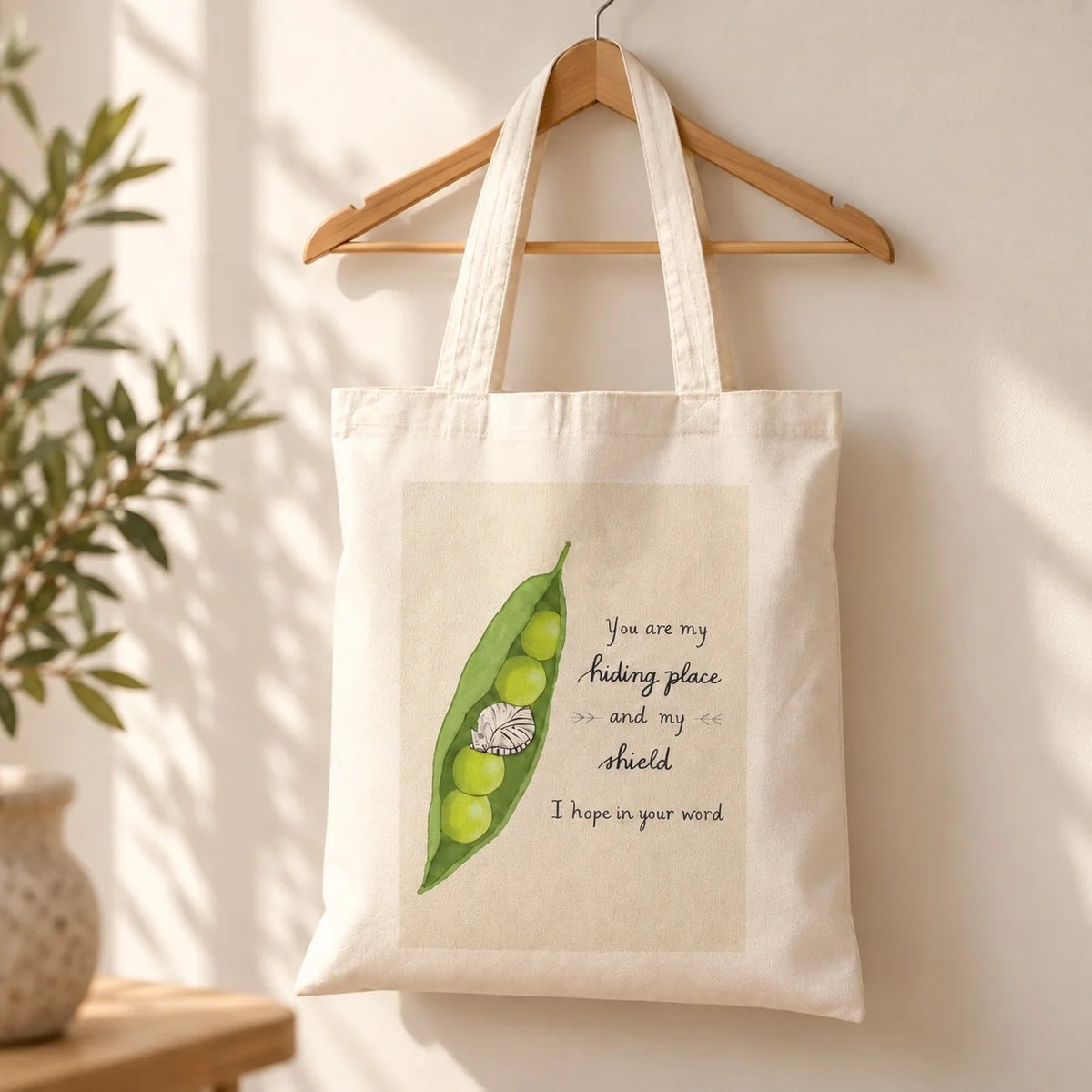 My Hiding Place Psalm 119:114 Christian tote bag with pea pod illustration on a wooden hanger in warm sunlight