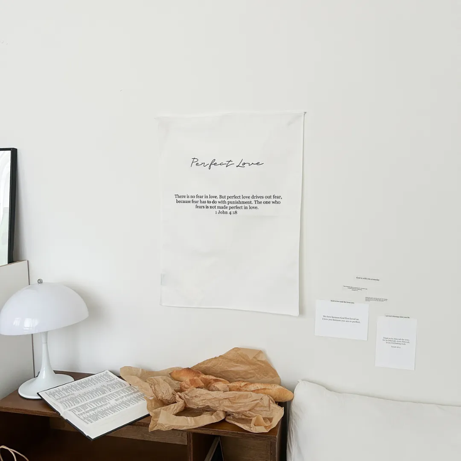 Minimalist wall tapestry with the words "Perfect Love" and 1 John 4:18 Bible verse, displayed above a cozy bedside table with bread, an open Bible, and soft lighting, inspiring faith and simple happiness.