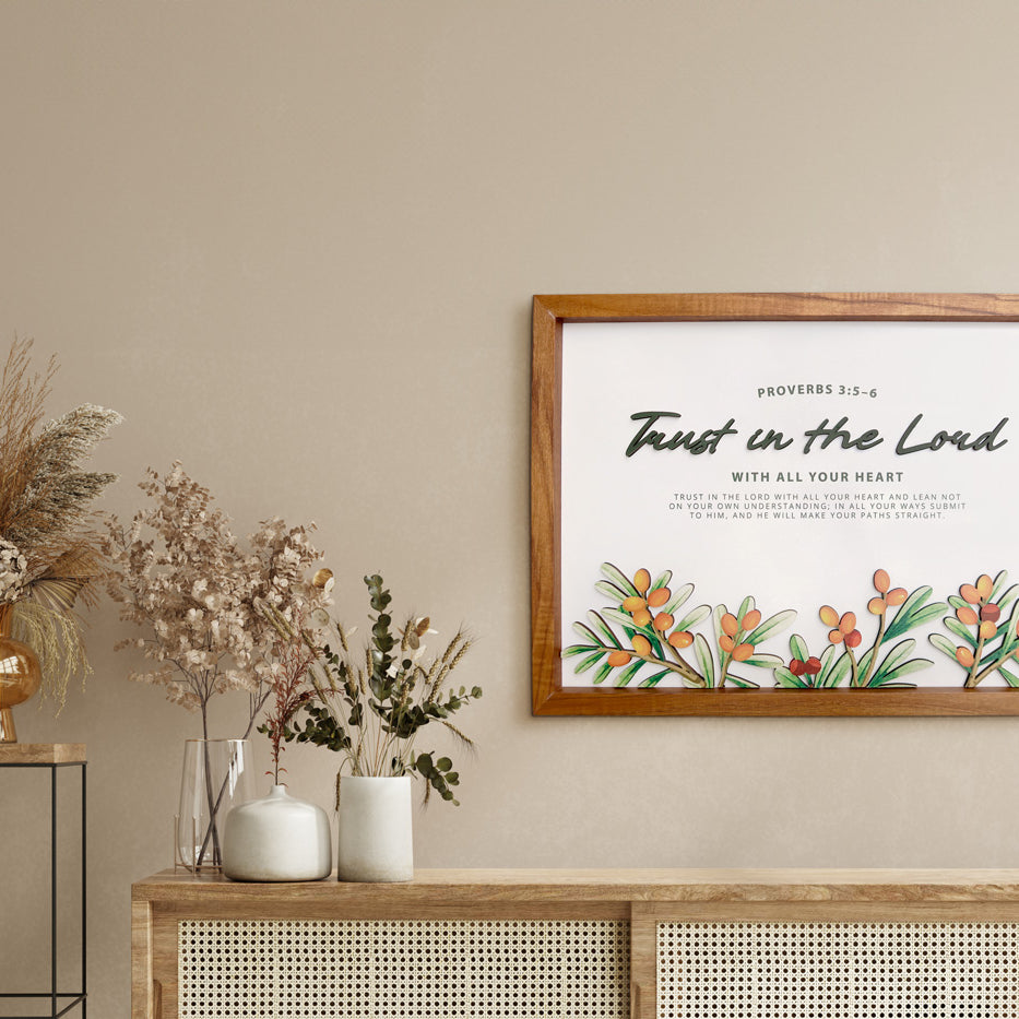A wooden-framed wall art featuring the Bible verse "Trust in the Lord" from Proverbs 3:5-6. The artwork is decorated with vibrant orange and green botanical designs. It is displayed in a neutral-toned room with a rattan cabinet adorned with dried flowers in vases, creating a modern yet rustic and serene ambiance ideal for Christian home decor.