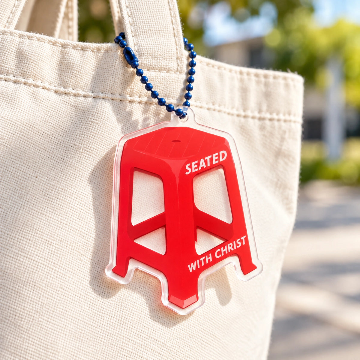 Red Chair Seated with Christ acrylic keychain hanging from a cream canvas tote bag outdoors.