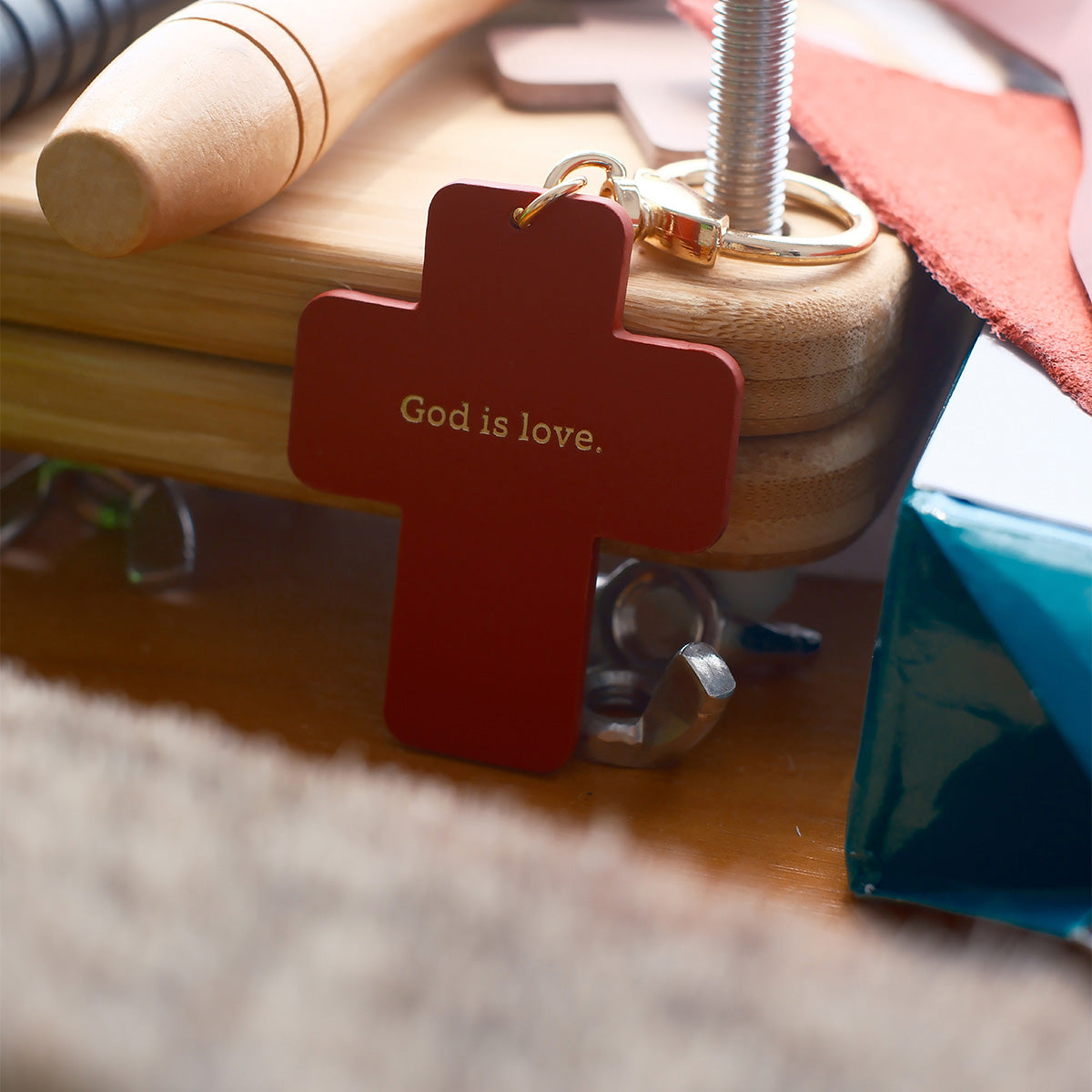 Red cross-shaped keychain with 'God is love' engraved, placed on a wooden desk with tools and accessories in the background.